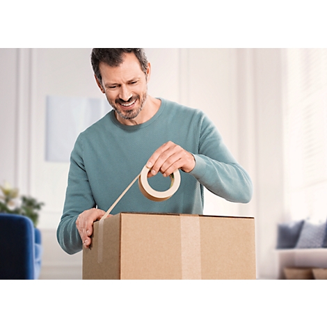 Een lachende man plakt een kartonnen doos dicht met plakband. Hij staat in een lichte kamer. Meubels zijn te zien op de achtergrond.