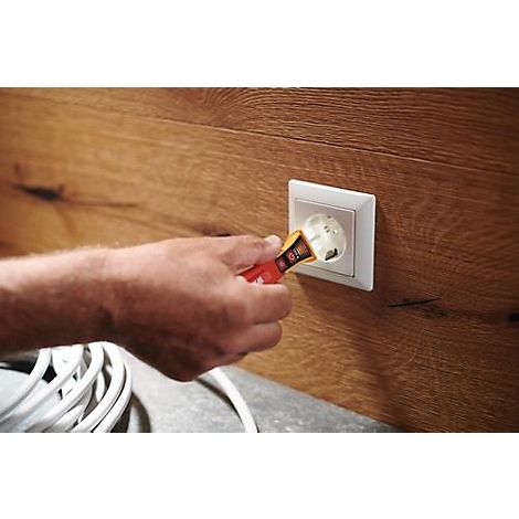 Une main teste une prise électrique avec un testeur de tension. Le fond montre du bois. Un câble est au premier plan.