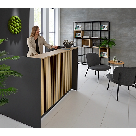 Eine Frau steht an einem Empfangstresen aus Holz und schwarzem Material. Im Hintergrund ist ein grünes Moosbild zu sehen.