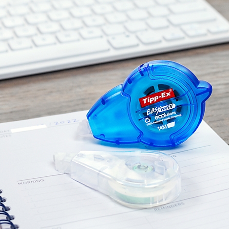 A blue Tipp-Ex Easyrefill next to a transparent spare part lie on a notebook. In the background, there's a keyboard.