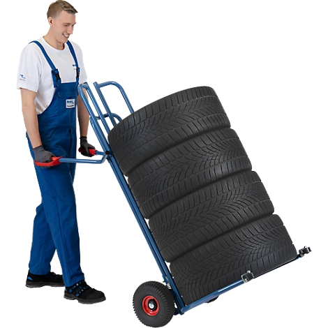 A man in overalls pushes a blue hand truck loaded with five car tires. He is smiling.
