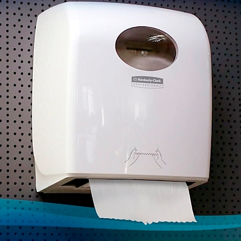 Dispensador de toallas de papel blanco sobre chapa perforada gris. Se saca papel. Logotipo 'Kimberly-Clark Professional'.