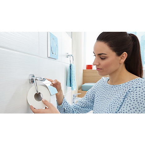 Woman in bathroom taking toilet paper from a holder. Behind her are tiles, a towel rack and a flush.