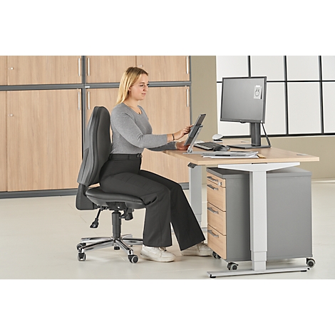 A woman sits at a desk working on a tablet. She wears a gray top and black pants.