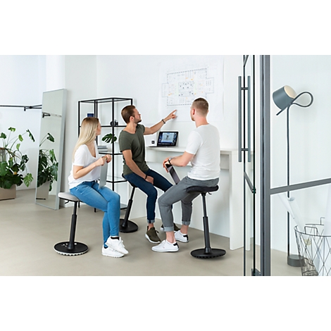 Three people sit on standing stools, looking at a drawing on the wall. One man points at the drawing as the others watch.