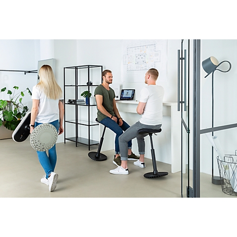 Three people in an office. Two are sitting on ergonomic stools, one person is walking with two stools.