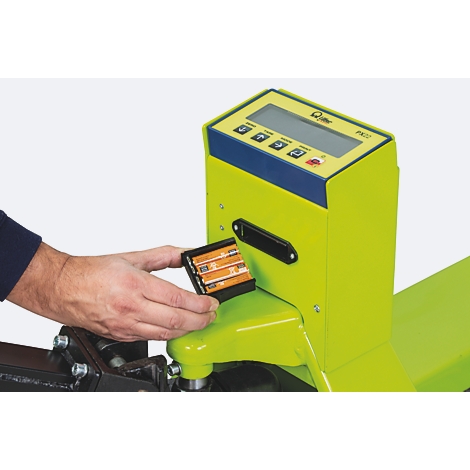 Primer plano de una persona insertando pilas en una báscula amarillo-verde. "PK22" es visible en la pantalla.