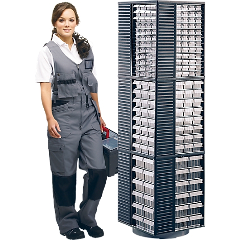 A woman in work clothes stands next to a black rotating rack with drawers. She is holding a toolbox.
