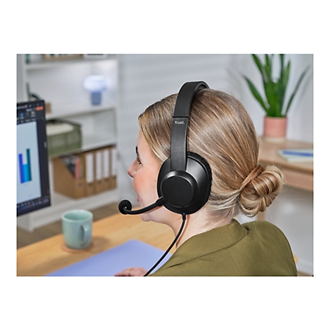 Eine Frau mit blondem Haar trägt ein schwarzes Headset. Im Hintergrund: Schreibtisch, Monitor mit Diagrammen und Büroregal.