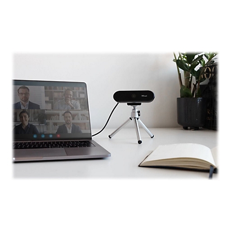 Schwarze Webcam auf Stativ vor Laptop mit Video-Konferenz, neben Pflanze und Notizbuch. Webcam mit Logo Trust.