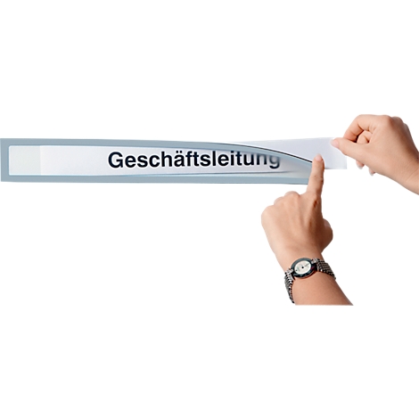 Two hands peel a strip labeled Geschäftsleitung from a sign. A wristwatch is visible on the wrist.