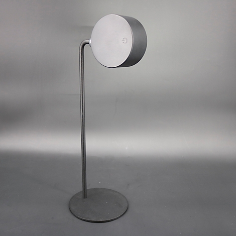 Eine metallene Stehlampe. Der runde Kopf ist an einem gebogenen Arm befestigt, der auf einer dünnen Stange steht. Auf dem Kopf ist ein Knopf.