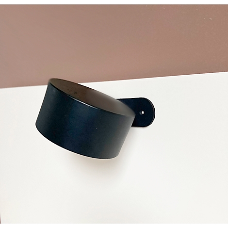 Schwarze Wandleuchte, zylindrisch, abgeschrägt, auf weißem Untergrund mit braunem Hintergrund. Flache Halterung zur Wandbefestigung.
