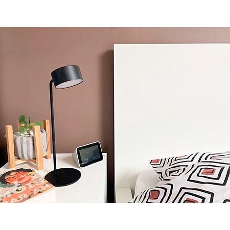 Schwarze Tischlampe auf Nachttisch neben einem weißen Bett, braune Wand. Auf dem Tisch ein Smart-Display, Pflanze und Buch.