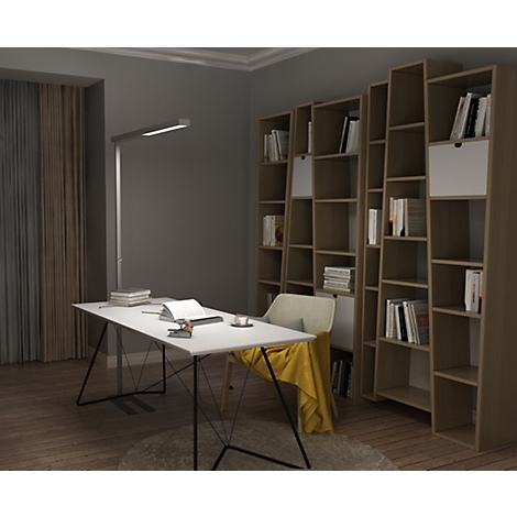 Ein moderner Schreibtisch mit Lampe und Stuhl, davor ein hohes Bücherregal in einem Wohnzimmer.