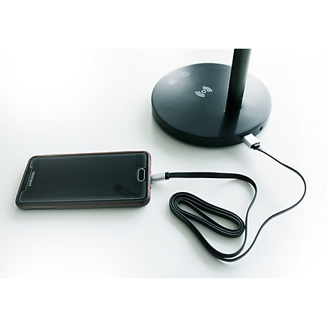Ein schwarzes Smartphone lädt per Kabel an einem schwarzen Standfuß einer Tischleuchte.