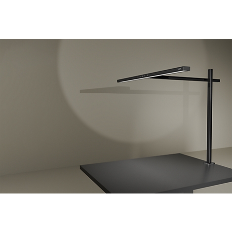Schwarze, minimalistische Schreibtischlampe leuchtet auf eine graue Wandfläche.