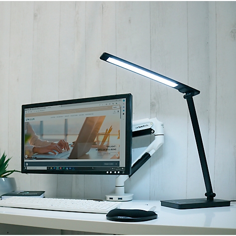 Schreibtisch mit Computer und schwarzer LED-Schreibtischlampe, die leuchtet. Auf dem Monitor ist eine Webseite zu sehen.