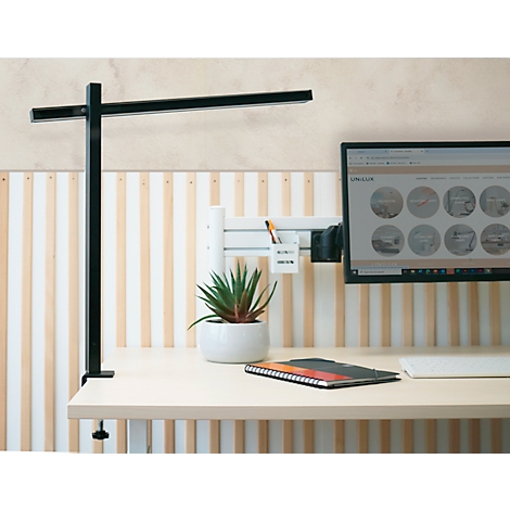 Schwarze Schreibtischlampe, Monitor, Pflanze, Notizblock auf einem hellen Schreibtisch. Im Hintergrund Holzvertäfelung.