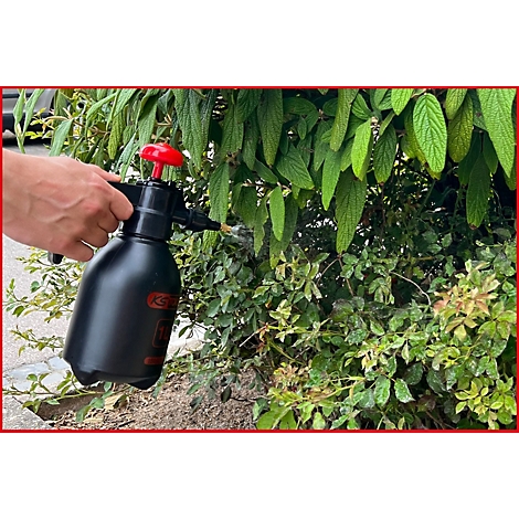Eine Person sprüht mit einer schwarzen Sprühflasche Pflanzen in einem Garten ein. Der rote Griff der Sprühflasche hebt sich ab.