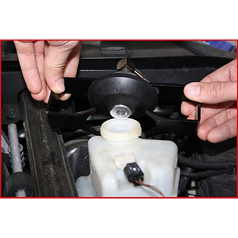 Hände halten ein schwarzes Werkzeug über einem weißen Behälter mit einer Öffnung. Im Hintergrund sind Autoteile zu sehen.