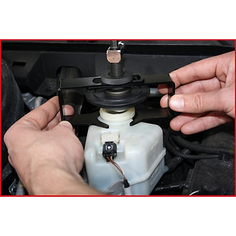 Hände halten ein schwarzes Werkzeug über einem Behälter im Motorraum eines Autos. Das Werkzeug hat eine Schraube und eine Halterung.