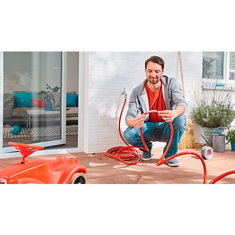 Man crouching, connecting red garden hose. In the background: house, toy car.