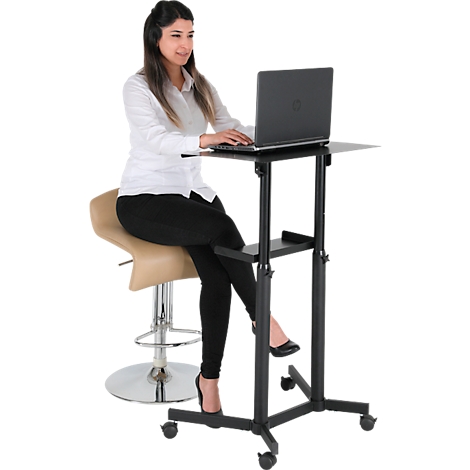 A woman sits at a height-adjustable standing desk with a laptop. She wears a white shirt and black trousers.