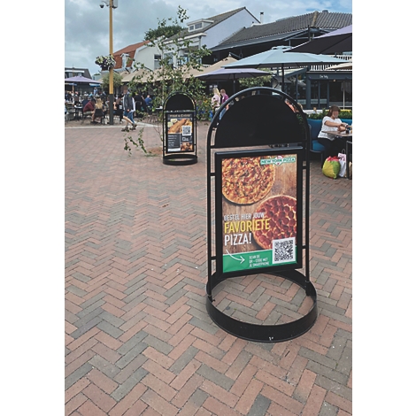 Deux panneaux de pizza sur une place pavée. En arrière-plan, des gens sont assis sous des parasols.
