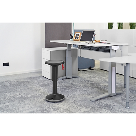 Office stool in front of height-adjustable desk. Desk with laptop, plants. Gray carpet, light walls.