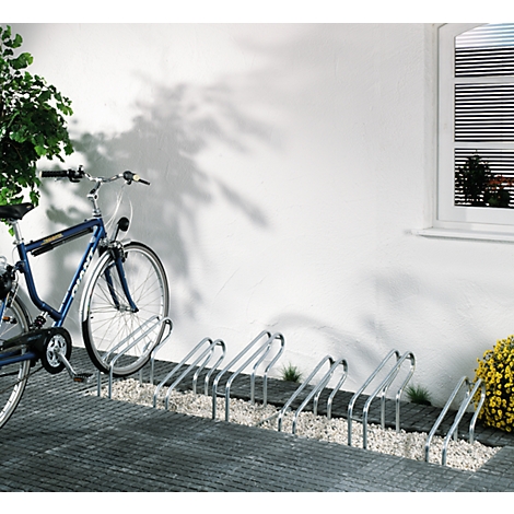Blue bicycle in a bike rack. The rack is mounted on a paved path in front of a white wall with a window.