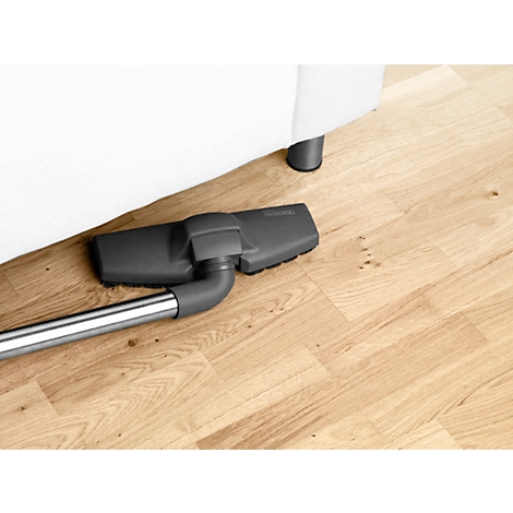 A vacuum cleaner head cleans the floor under a white sofa. The floor has a light wood pattern.