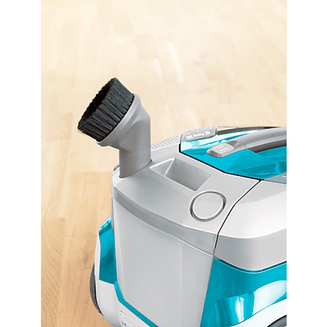 Detail of a vacuum cleaner with a brush. The vacuum cleaner has a light gray base, blue elements and a wooden floor as a background.