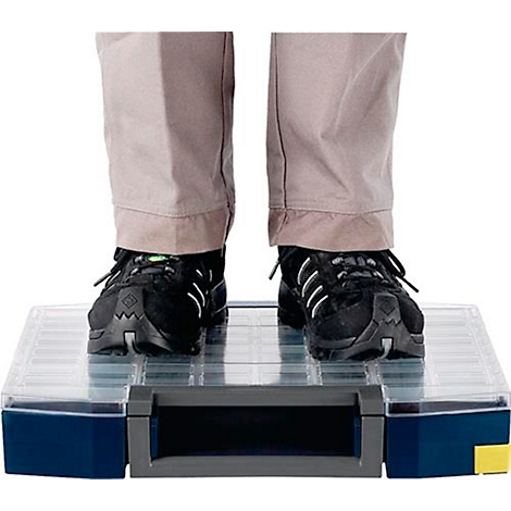 Une personne se tient debout sur une boîte à outils transparente. Chaussures noires, pantalon beige. Boîte à outils avec des accents bleus et jaunes.