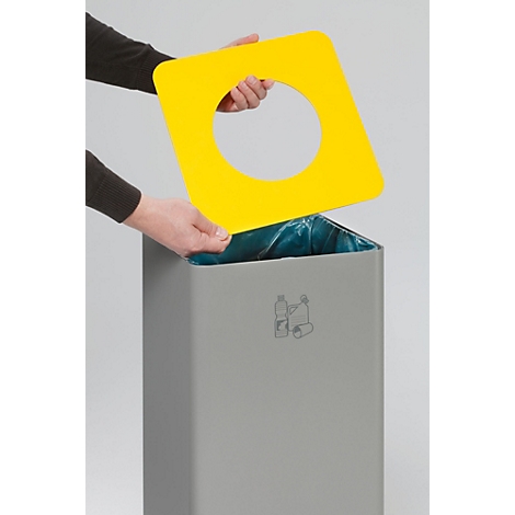 Een persoon houdt een geel vierkant deksel met een rond gat boven een grijze vuilnisbak.