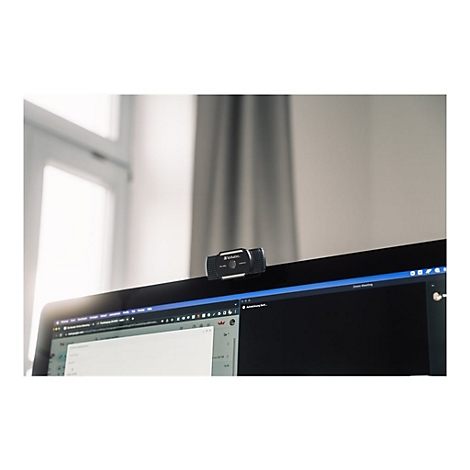 Schwarze Webcam auf einem Monitor. Im Hintergrund sind ein Fenster und VorhÃ¤nge zu sehen.