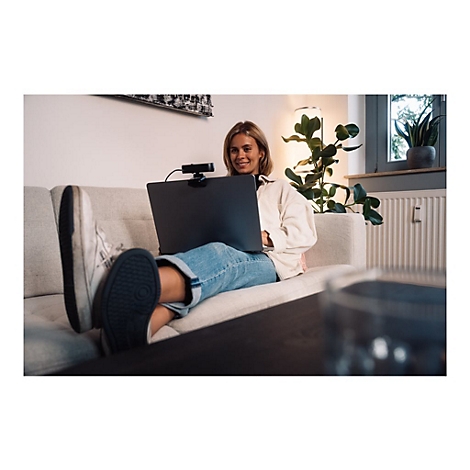 Eine lÃ¤chelnde Frau sitzt entspannt auf einem Sofa mit Laptop und Webcam.