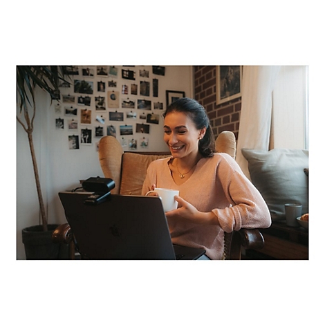 Eine lÃ¤chelnde Frau sitzt vor einem Laptop und hÃ¤lt eine Tasse. Im Hintergrund sind Fotos an der Wand und eine Topfpflanze zu sehen.