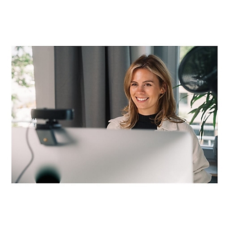 Eine lÃ¤chelnde Frau sitzt vor einem Computer mit Webcam. Helle Kleidung, blondes Haar, Vorhang im Hintergrund.