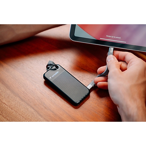 Eine Hand schließt eine kleine schwarze Verbatim-SSD mit kurzem USB-C-Kabel an ein Tablet auf einem Holztisch an.