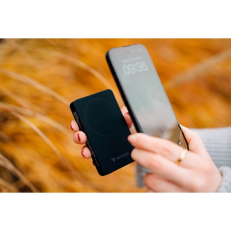 Hand hält ein schwarzes MagSafe-Ladegerät neben ein Smartphone vor unscharfem herbstlichem Hintergrund.