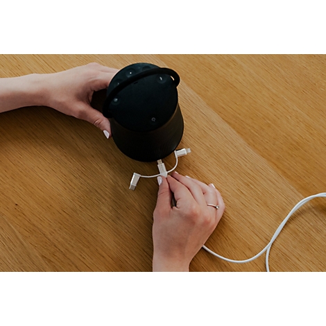 Hände halten einen schwarzen Smart-Speaker und stecken ein weißes Ladekabel in den Anschluss.