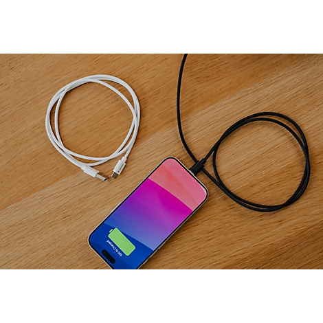 Smartphone lädt mit schwarzem Kabel auf einer Holzoberfläche, daneben ein weißes Ladekabel.