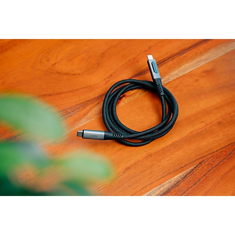 Ein aufgerolltes schwarzes USB-C-Kabel liegt auf einem hölzernen Tisch, unscharfe grüne Pflanzen im Vordergrund.