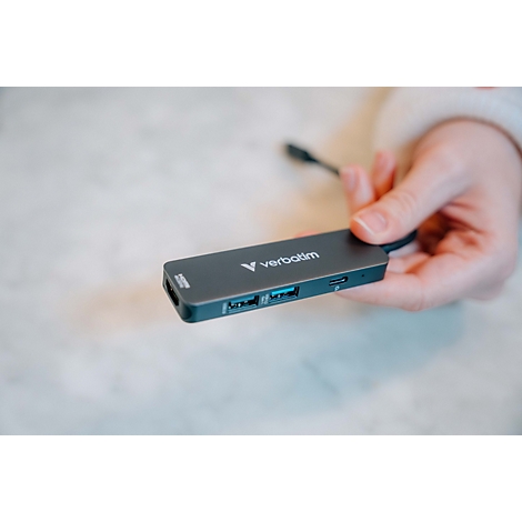 Hand hält einen grauen Verbatim USB-C-Hub mit verschiedenen Anschlüssen über einem hellen Marmortisch.