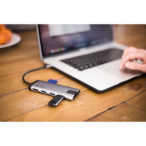 Zilveren USB-C hub op een houten tafel naast een laptop. Een hand bedient het toetsenbord. Er is een SD-kaartlezer geplaatst.