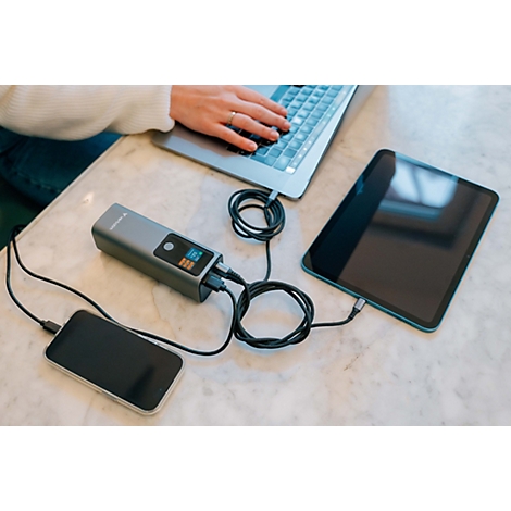 Eine Powerbank lädt gleichzeitig einen Laptop, ein Tablet und ein Smartphone auf einem hellen Tisch.