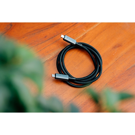 Aufgerolltes schwarzes USB-C-Kabel auf einem hölzernen Untergrund mit unscharfen grünen Pflanzen im Vordergrund.