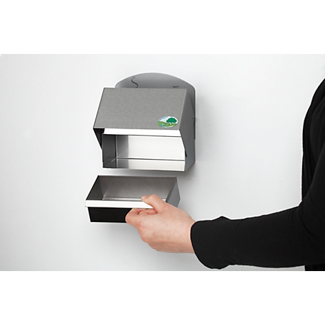 A person opens a metal paper towel dispenser on a white wall. The dispenser has a green logo.