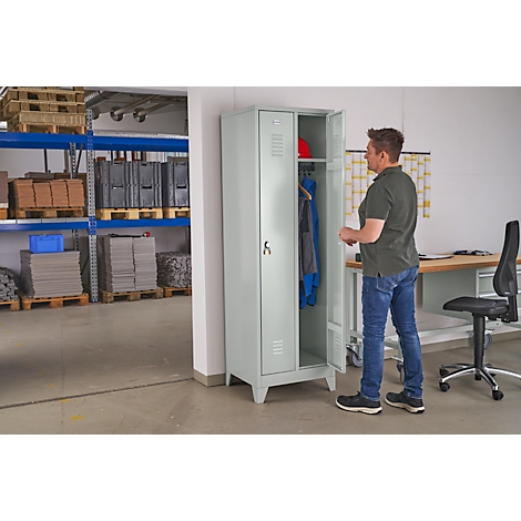 A man stands in front of an open, light gray locker. Clothing and a helmet are visible inside. The man is wearing jeans and a shirt.